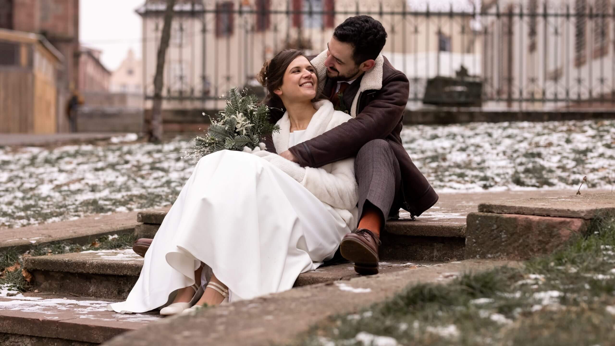 Photos de couple : Après avoir dis oui à l'église, les mariés se prennent un petit moment à deux pour une séance photo. Les mariés sont assis sur les marches derrière l'église. La mariée est entre les jambes du mariés et se regardent.