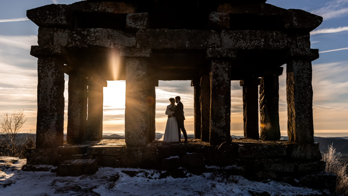 Un temple d'amour : Deux mariés en tenues, s'enlacent dans le temple du Donon situé au sur un sommet des Vosges dans le Grand - Est. Le soleil est en arrière plan et se couche entre les colonnes du temple.