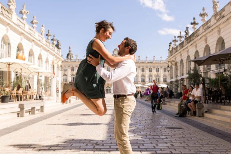 Saut vers l'avenir : Séance engagement des mariés sur la place Stanislas à Nancy. Les mariés sont face à face. La mariée saute en l'air et le marié l'aide en la soutenant par les hanches.