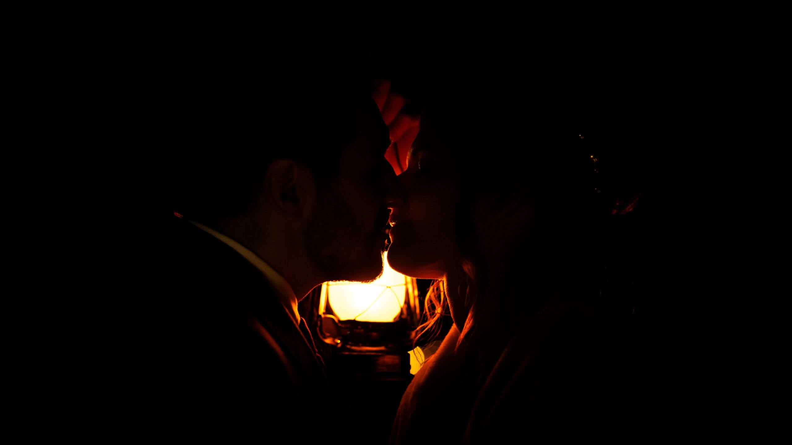 Une lumière douce : Deux mariés en tenues dans le noir éclairés par une lampe à pétrole. La photo est pris en cadrage serré et la lueur de la lampe se reflète sur leur visage pendant qu'ils s'embrassent.