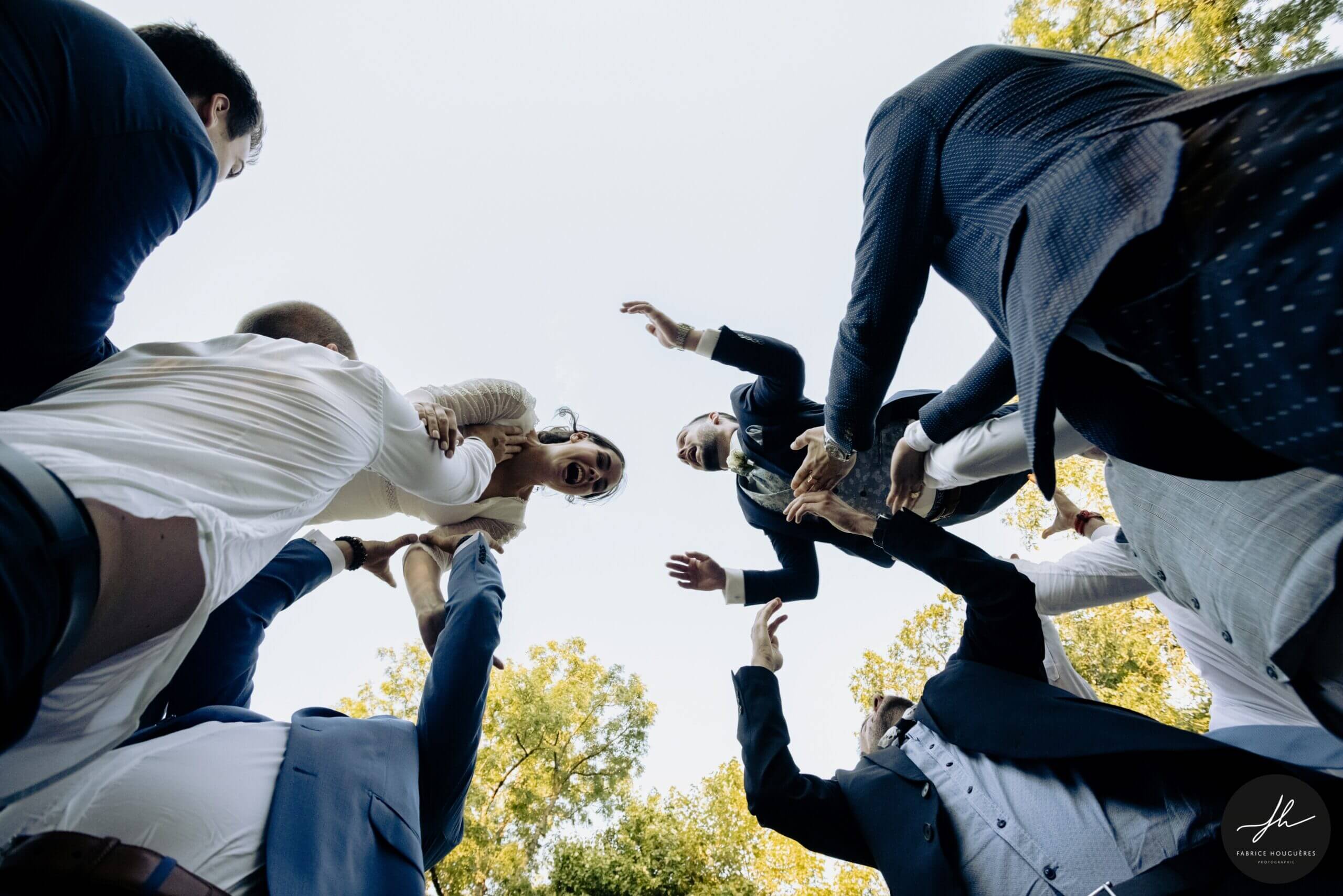 Prêt au décollage? La photo est prise en contre plongée. Les invités envoient les mariés en l'air face à face. Les mariés rigolent.